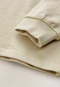 Hellbeige Langarmshirt mit gerippter Textur, ausgestattet mit umgeschlagenen Manschetten und einem flachen Saum, das ein einfaches, glattes Design zeigt.