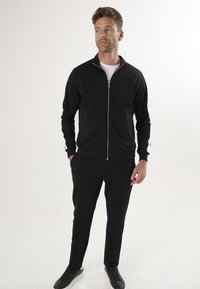 Schwarze Zip-Jacke mit Rippbündchen und weißen Akzenten an den Ärmeln, kombiniert mit schwarzen Anzughosen und schwarzen Sneakers.
