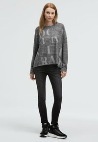 Grauer Pullover mit großen, hellgrauen Textmustern; kombiniert mit schwarzen Skinny-Jeans und schwarzen, fellgefütterten Sportschuhen.