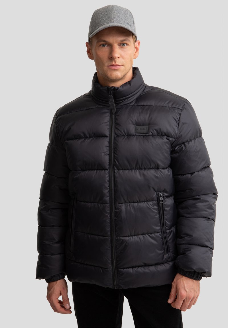 Melnā pūkainā jaka ar horizontālu quilted dizainu un augstu apkakli, aprīkota ar zipa sānu kabatām un zīmola etiķeti uz krūtīm.