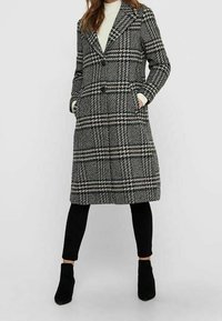 Dvojřadý kabát s černobílým houndstooth vzorem, který má velké špičaté klopy, boční kapsy a délku ke kolenům.