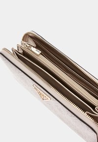 Beige plånbok med en texturerad yta, gyllene detaljer och flera fack. Har en dragkedja och en präglad logotyp på framsidan.