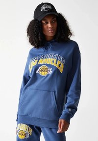 Sweat à capuche bleu avec le logo "Los Angeles Lakers" en blanc et jaune. Comprend une poche avant et des poignets côtelés. Accompagné d'un pantalon assorti.