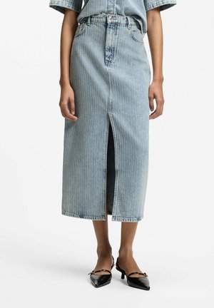 Kvinde iført en lyseblå denimnederdel til midt på læggen med en frontslids og sorte lavhælede sko med spidse tæer.