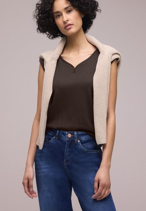 Jeune femme aux cheveux bouclés portant un haut marron à manches courtes, un jean bleu et un pull en maille beige drapé sur les épaules, sur un fond uni.