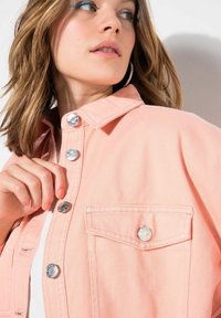 Chaqueta de mezclilla melocotón con cierre de botones en la parte delantera, herrajes de metal plateado, cuello y un bolsillo en el pecho. La tela tiene una textura suave.