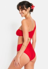 Bikini rouge à une épaule avec un devant froncé, bas taille haute et détail découpé. Tissu lisse et extensible avec une finition brillante.