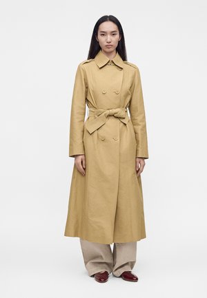 Femme portant un long trench-coat beige à double boutonnage, noué à la taille, pantalon beige large et chaussures rouge foncé, debout devant un fond blanc uni.