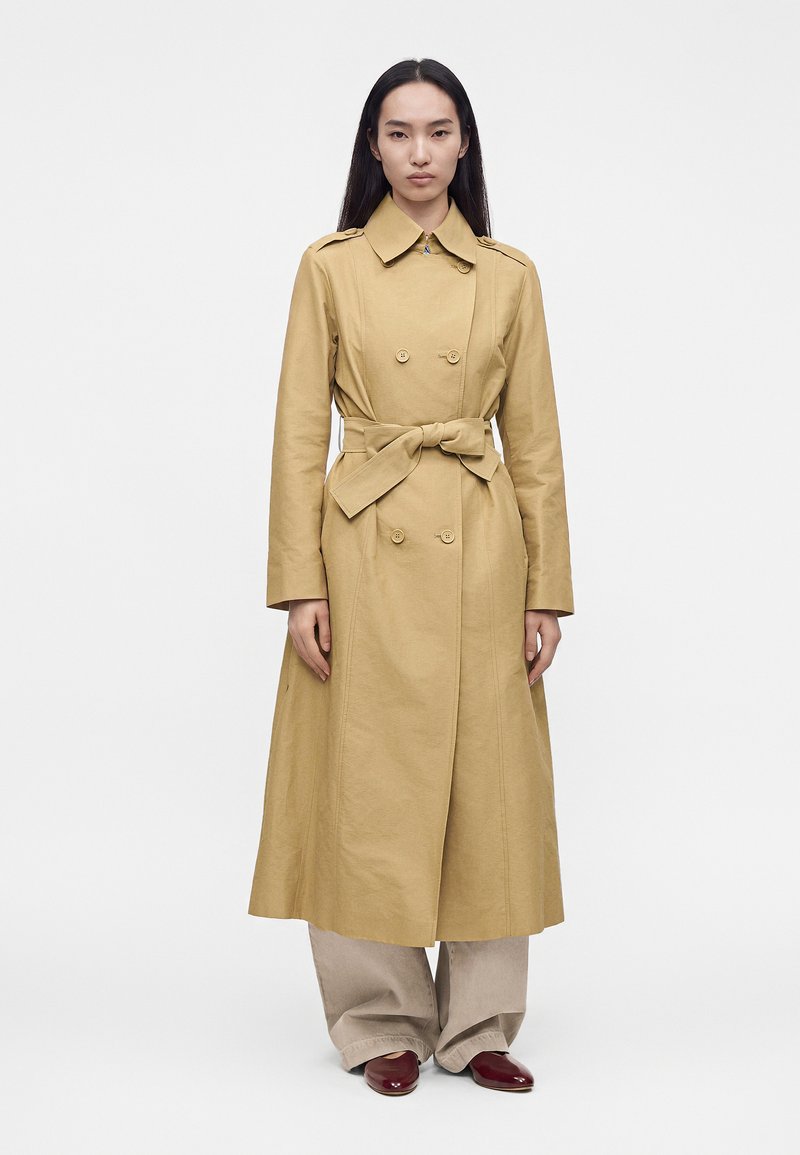 Femme portant un long trench-coat beige à double boutonnage, noué à la taille, pantalon beige large et chaussures rouge foncé, debout devant un fond blanc uni.