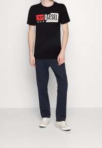 Czarna bawełniana koszulka z białym i czerwonym logo Diesel, zestawiona z granatowymi spodniami bawełnianymi i białymi trampkami. Prosty, casualowy design.