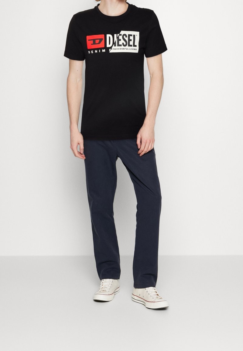 Czarna bawełniana koszulka z białym i czerwonym logo Diesel, zestawiona z granatowymi spodniami bawełnianymi i białymi trampkami. Prosty, casualowy design.