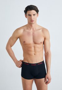 Schwarze Boxershorts mit enganliegendem Schnitt, ausgestattet mit einem roten elastischen Bund, der mit "HUGO" gebrandet ist. Glatte Textur, entworfen für Komfort und Unterstützung.