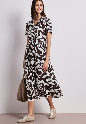 Frau steht lächelnd da, trägt ein Midi-Kleid mit abstraktem schwarz-weißem Druck und Knöpfen sowie beige Sneakers und hält eine taupefarbene Tasche.