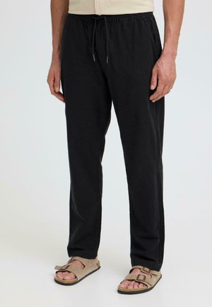 Mann trägt schwarze Kordelhose und beige Sandalen mit zwei Schnallen, steht vor einem schlichten hellen Hintergrund.