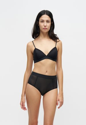 Femme aux longs cheveux foncés portant un bralette noir et une lingerie résille noire taille haute, debout sur un fond clair uni.