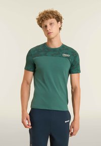 T-shirt vert à manches courtes avec un panneau supérieur à motif camouflage et un logo sur l'épaule gauche. Texture lisse, design ajusté.