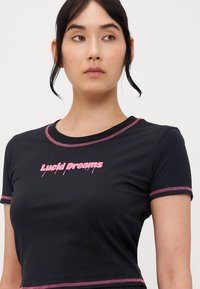 Černé tričko s krátkým rukávem, na kterém je růžová grafika "Lucid Dreams", s kontrastním prošíváním kolem límce a rukávů, vyrobené z měkké látky.