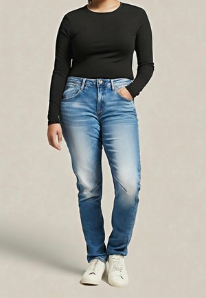 Person wearing a fitted black long-sleeve top, blue faded jeans with one hand in pocket, and white sneakers, standing on neutral background.