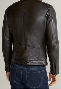 Hnědá kožená motocyklová bunda s přiléhavým střihem, prošívanými ramenními detaily a zipy na manžetách. Nošena přes modré džínové kalhoty.