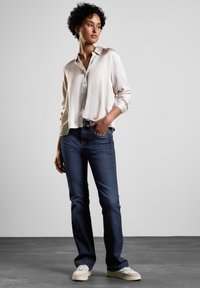Chemise en satin beige clair avec un col, associée à un jean en denim bleu foncé. Baskets blanches, sur un fond neutre.