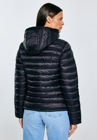 Vrouw draagt een zwarte gewatteerde jas met capuchon en lichtblauwe spijkerbroek, staand met haar rug naar de camera tegen een effen grijze achtergrond.