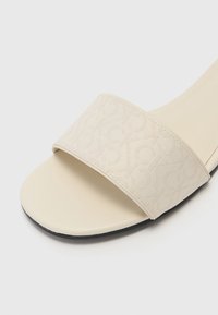 Sandale à glissière beige avec une large sangle en tissu arborant un motif de monogramme subtil. Semelle douce avec une forme arrondie et flexible, et une surface texturée.