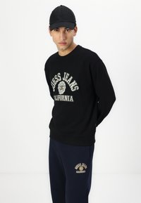 Sudadera negra con el logo "Guess Jeans California"; cuenta con puños y dobladillo acanalados. Combinada con pantalones de chándal azul marino que tienen un logo a juego. Gorra negra.