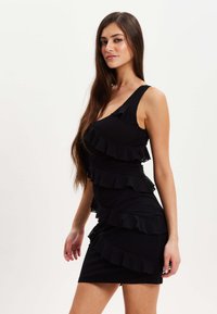 Black mini dress with layered ruffle design, fitted silhouette, and wide shoulder straps. Smooth fabric with a textured finish.