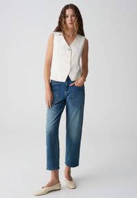 Weißes ärmelloses Oberteil mit V-Ausschnitt und fünf Knöpfen, kombiniert mit blauen hochtaillierten Jeans und cremefarbenen Slippern.