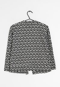 Svartvit mönstrad jacka upphängd på en galge med en geometrisk hexagonal design och öppen front, som visar en texturerad tyg.