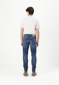 Jeans in denim blu da uomo con una vestibilità slim, cinque tasche e un'etichetta in pelle color beige sul retro. Abbinati a una maglietta grigia chiara e calzature beige.