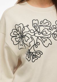 Ljus beige sweatshirt med ett framträdande svart blommigt broderi på framsidan. Enkel halsringning och avslappnad passform.