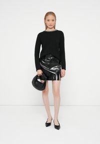 Mulher a usar um suéter preto de malha em corda com decote de pérolas, uma saia mini de couro verniz preto, sapatos rasos pretos de bico e a segurar uma bolsa preta.