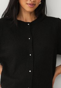 Cardigan nero a maniche corte realizzato in morbido tessuto, con scollatura rotonda e cinque bottoni lucidi lungo il davanti.