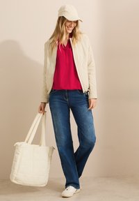 Veste bomber crème clair sur un haut fuchsia, associée à un jean bleu. Tient un sac cabas matelassé crème et porte une casquette beige. Baskets blanches.