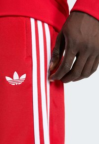 Červené športové nohavice s tromi bielymi zvislými pruhmi pozdĺž strán a bielym logom na dolnej časti nohy. Materiál pôsobí hladko a mäkko.