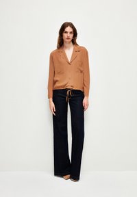 Braunes, langärmliges Hemd mit Kordelzug an der Taille und zwei Brusttaschen, kombiniert mit dunklen, weit geschnittenen Jeans und beigen Heels. Glatte Textur.