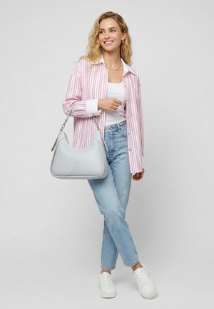 Naine, kes kannab roosade triipudega särk, valget tankitoppi, helesiniseid teksapükse, valgeid tossusid, hoides helehallit õlakotti, naeratab ja vaatab küljele.