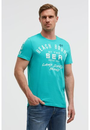 Mann trägt ein türkisfarbenes T-Shirt mit der Aufschrift "Beach Bound at the Sea, Barefoot Vibes, Camp David Brand" und blaue Jeans, die Hand in der Tasche.
