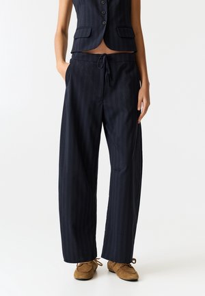 Femme portant un pantalon large à fines rayures bleu marine, un gilet boutonné assorti et des mocassins en daim marron, debout les mains dans les poches.