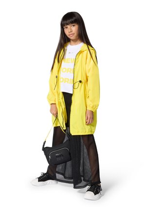 Gele lichtgewicht jas met trekkoord, wit t-shirt met print, zwarte rok van mesh, zwarte crossbody tas en zwarte sneakers met witte zolen.