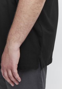 Černé bavlněné tričko s kulatým výstřihem, postranními rozparky a krátkými rukávy, vystavené na jednobarevném pozadí s rukou odpočívající poblíž.