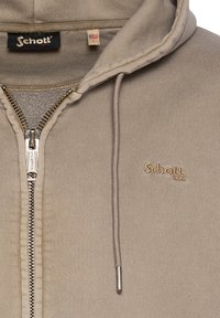 Beige zip-up hættetrøje med en struktureret overflade, der har en frontlynlås, snorehætte og et broderet logo på brystet.