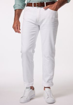 Homme portant un pantalon blanc retroussé, des baskets blanches, une ceinture marron et une chemise manches longues vert clair, une main dans la poche.