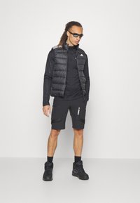 Crna punjena prsluk preko crne majice s dugim rukavima, u kombinaciji s crnim kratkim hlačama s džepovima i božnim logom, završeno s crnim tenisicama.