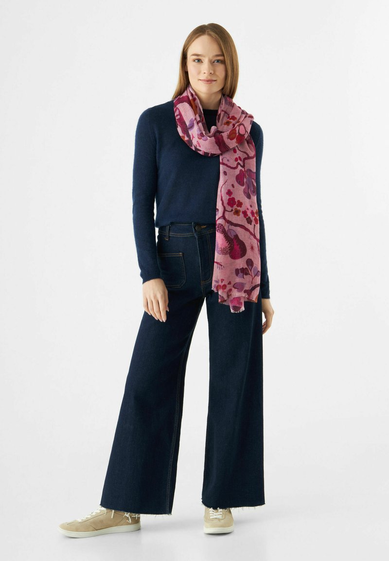 Écharpe rose à motif floral enroulée autour d'un pull bleu marine ; un pantalon large en denim foncé taille haute et des sneakers beiges complètent le look.