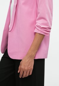 Ružový blejzer s hladkým materiálom, s klopovým golierom a pomačkanými rukávmi. Spárovaný s upravenými čiernymi nohavicami, ktoré majú jednoduchý dizajn.
