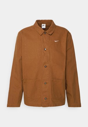 Ruda medvilninė striukė su apykakle, priekiniais mygtukais, dviem didelėmis kišenėmis ir maža balta Nike logo viršutiniame kairiajame krūtinės kampe.