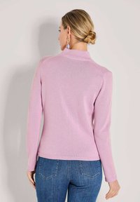 Frau mit blonden Haaren zu einem Dutt gebunden, trägt einen helllila Langarmpullover und blaue Jeans, steht mit dem Rücken zur Kamera.