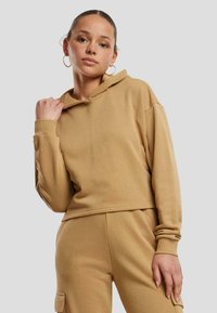 Sudadera recortada de color beige hecha de tela suave, con capucha ajustable, mangas largas y puños de canalé, llevada con pantalones a juego. Textura suave.